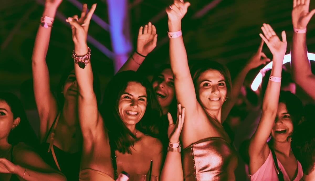 Two women smiling with hands raised during a terrace club party at night.