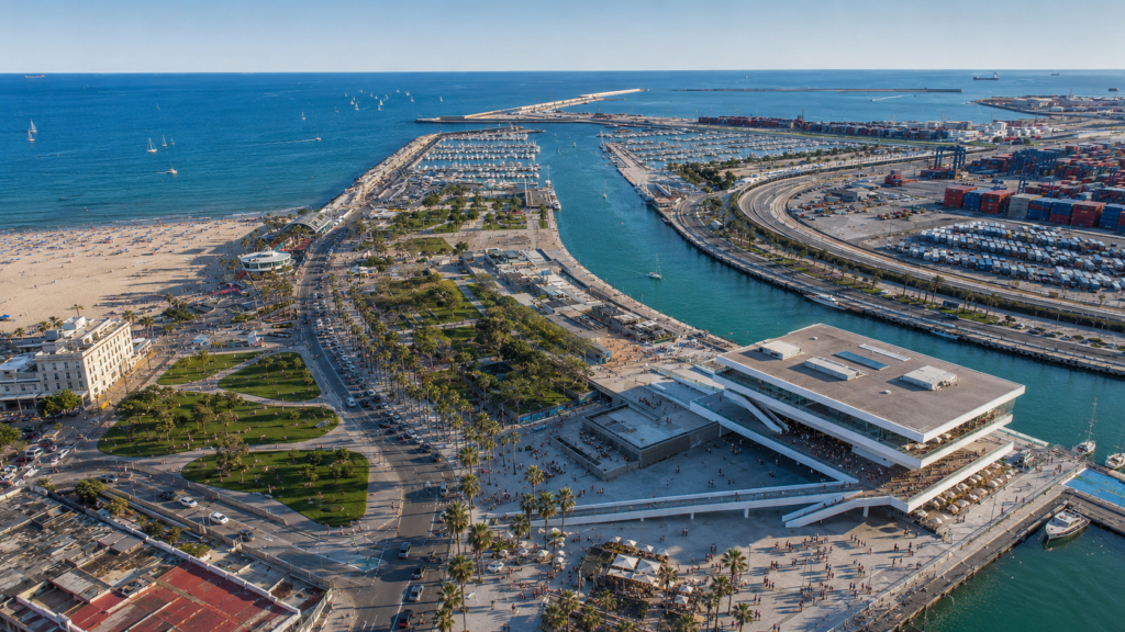 Vista aérea del Puerto de Valencia y la Marina de Valencia de día, con el edificio Veles e Vents, zonas de ocio, playa, paseo marítimo y locales junto al mar.
